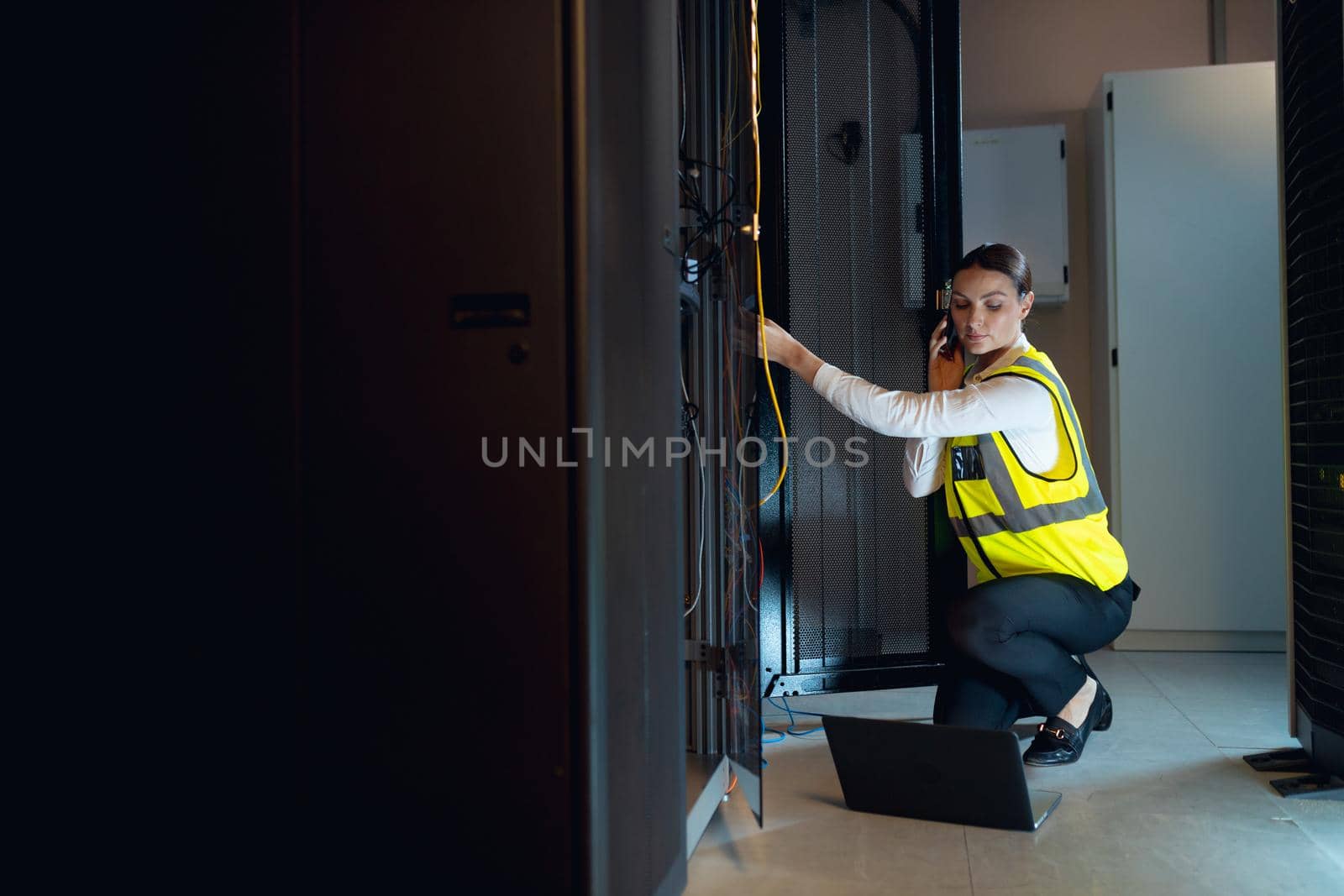 Caucasian female engineer with laptop talking on smartphone while inspecting in computer server room. database server management and maintenance concept