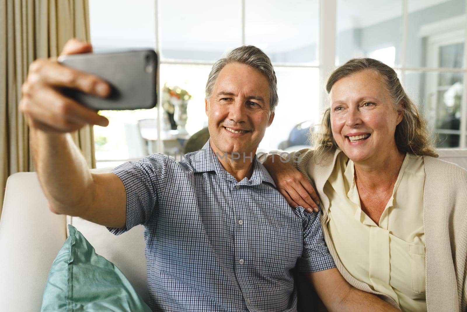 Happy senior caucasian couple in living room, sitting on sofa, taking selfies and smiling. retirement lifestyle, spending time at home.