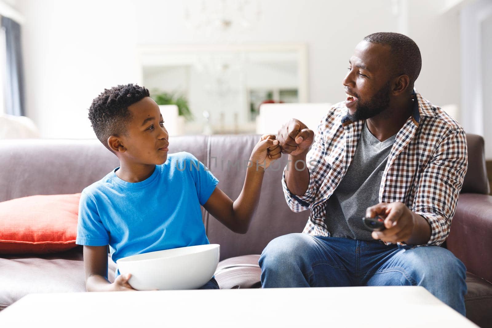 Happy african american father and son sitting on sofa, fist bumping watching tv. family spending time together at home.