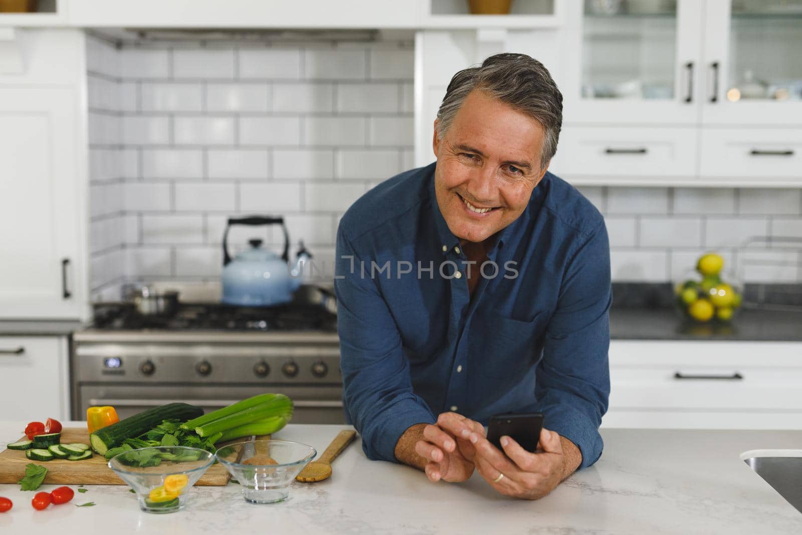 Portrait of senior caucasian man in modern kitchen, using smartphone, looking to camera by Wavebreakmedia