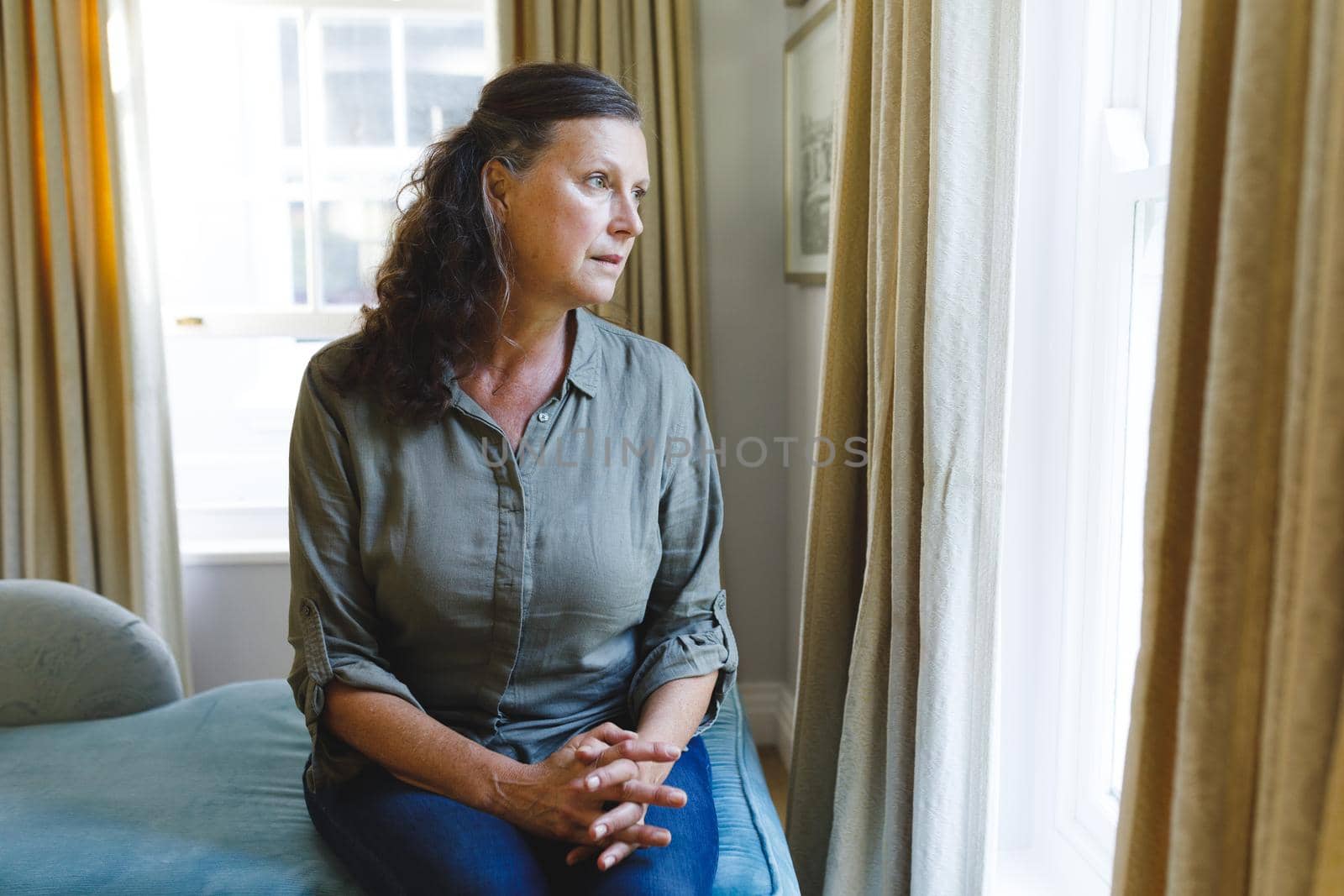 Thoughtful senior caucasian woman sitting on sofa and looking through window. retirement lifestyle, spending time at home.