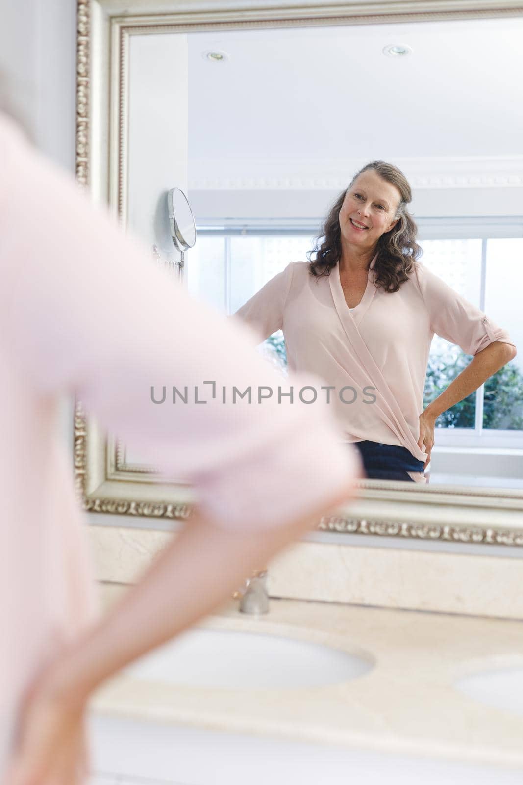 Happy senior caucasian woman in bathroom, looking in mirror and smiling. senior caucasian woman in bathroom, looking at her face in mirror. retirement lifestyle and the aging process.