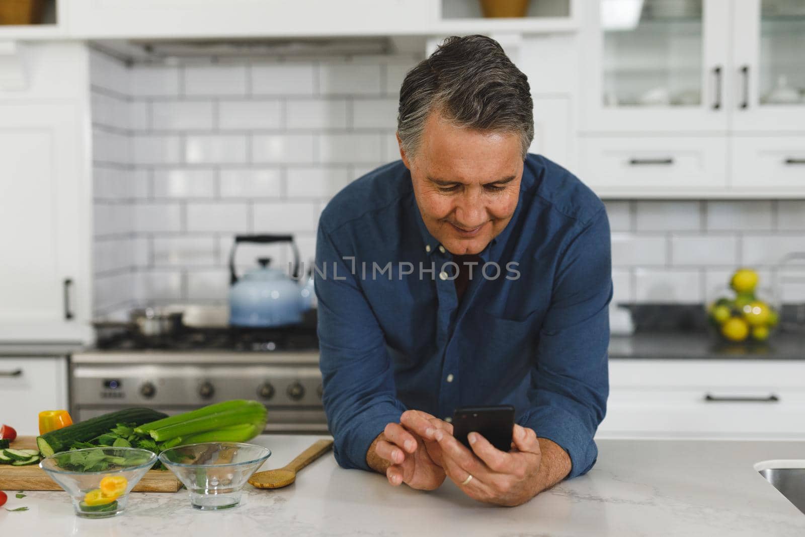 Smiling senior caucasian man in modern kitchen, using smartphone. retirement lifestyle, spending time at home.