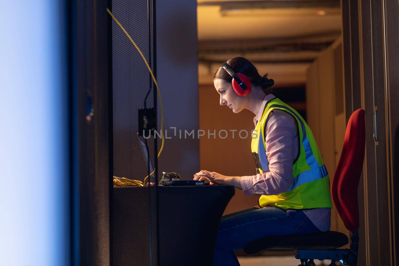 Caucasian female engineer wearing ear plugs using a laptop in computer server room by Wavebreakmedia