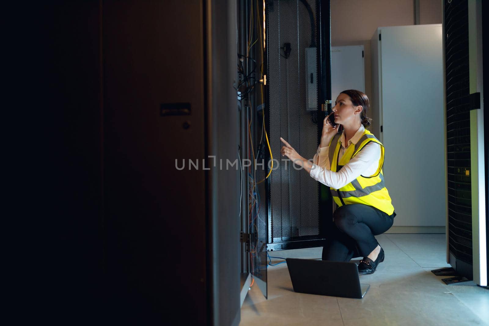 Caucasian female engineer with laptop talking on smartphone while inspecting in computer server room. database server management and maintenance concept