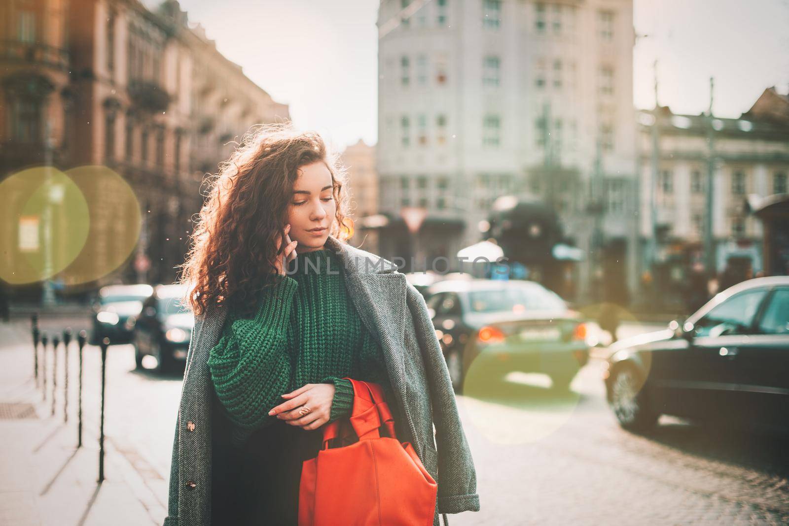 A woman on the street uses a mobile phone. online shopping. use of mobile applications. beautiful young woman with long curly dark hair in a casual coat, trendy green sweater and red handbag