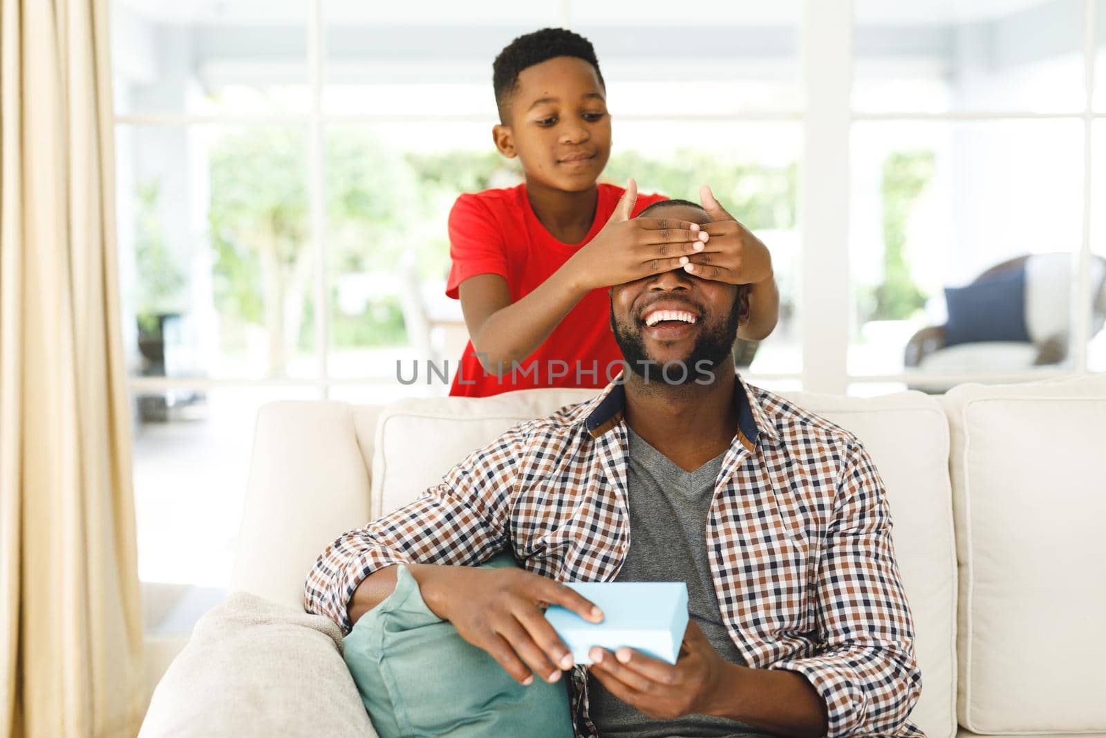 Smiling african american father opening gift from his son while he covers his eyes in living room. family spending time at home, father son relationship.