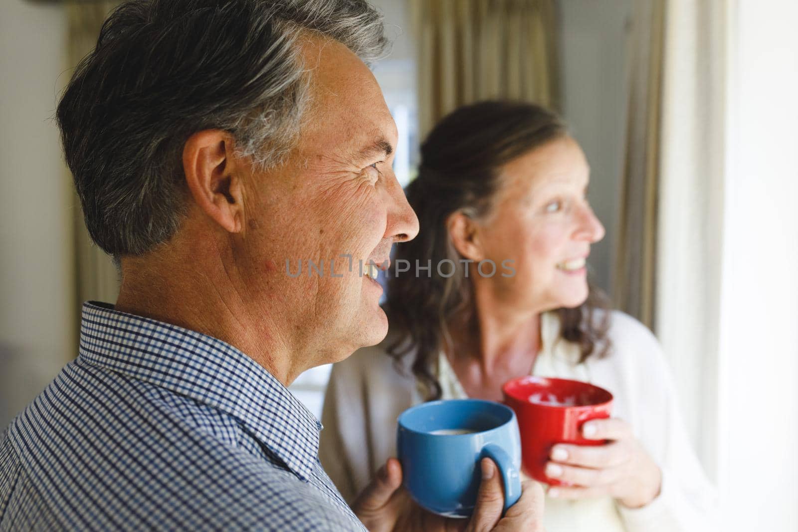 Happy senior caucasian couple standing next to window, holding cups. retirement lifestyle, spending time at home.