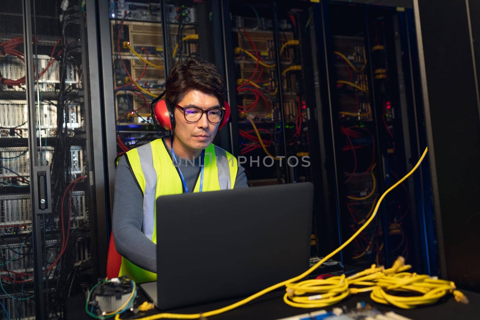 Asian male engineer wearing ear plugs using a laptop in computer server room. database server management and maintenance concept