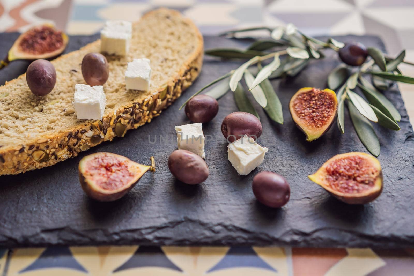 Green and black olives with loaf of fresh bread, feta cheese and young olives branch on olive wood chopping board over dark background.