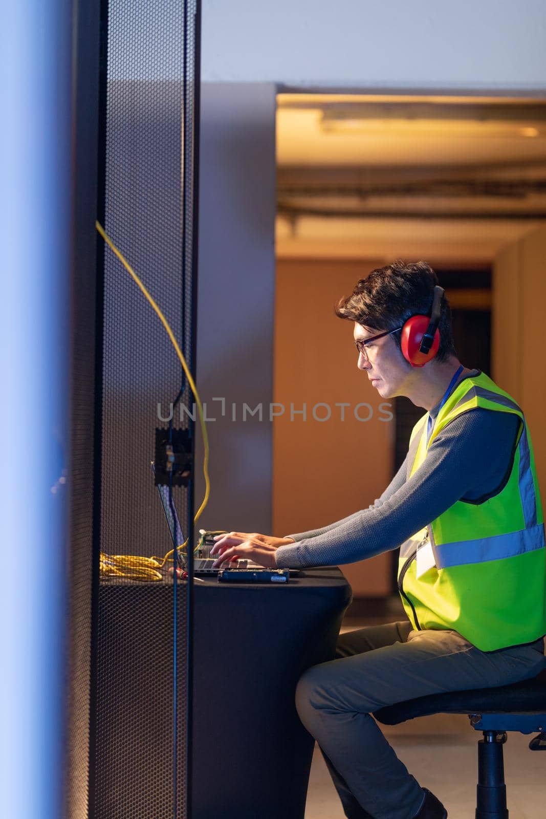 Asian male engineer wearing ear plugs using a laptop in computer server room by Wavebreakmedia