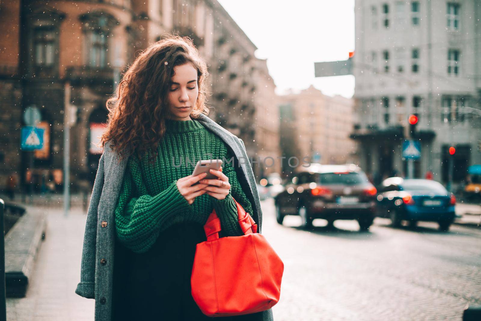 A woman on the street uses a mobile phone. online shopping. use of mobile applications. beautiful young woman with long curly dark hair in a casual coat, trendy green sweater and red handbag