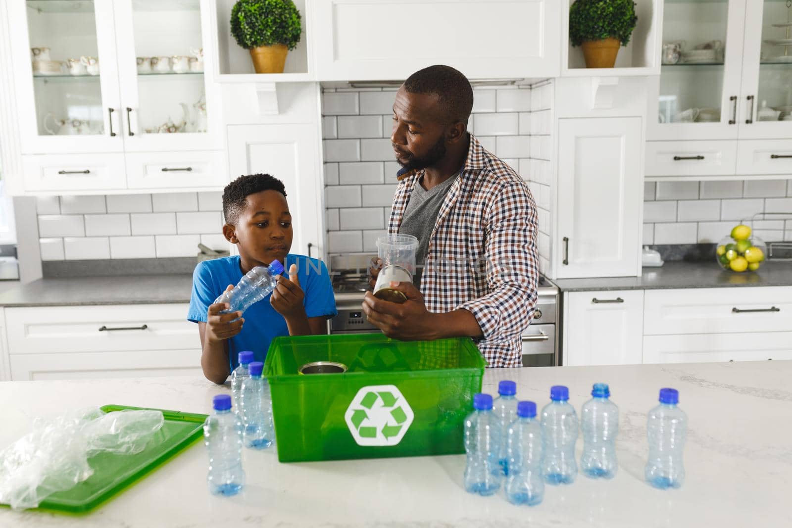 Happy african american father and son in kitchen talking and sorting plastic waste for recycling. family spending time at home.