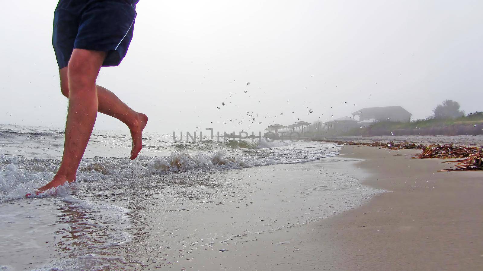 Splashes from the waves are flying around. sand and waves under the feet of a man running in shorts. Barefoot morning jogging on the beach. The feet of a man running along a sea beach in foggy weather.