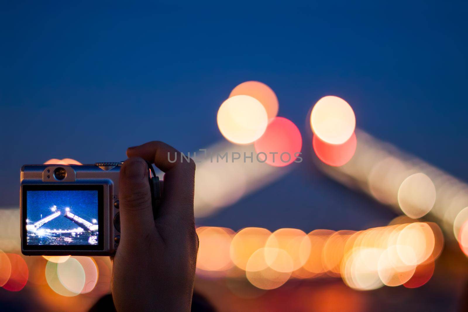 Hand with a silver compact camera shooting night scene, selective focus on a camera with lens blur on background.