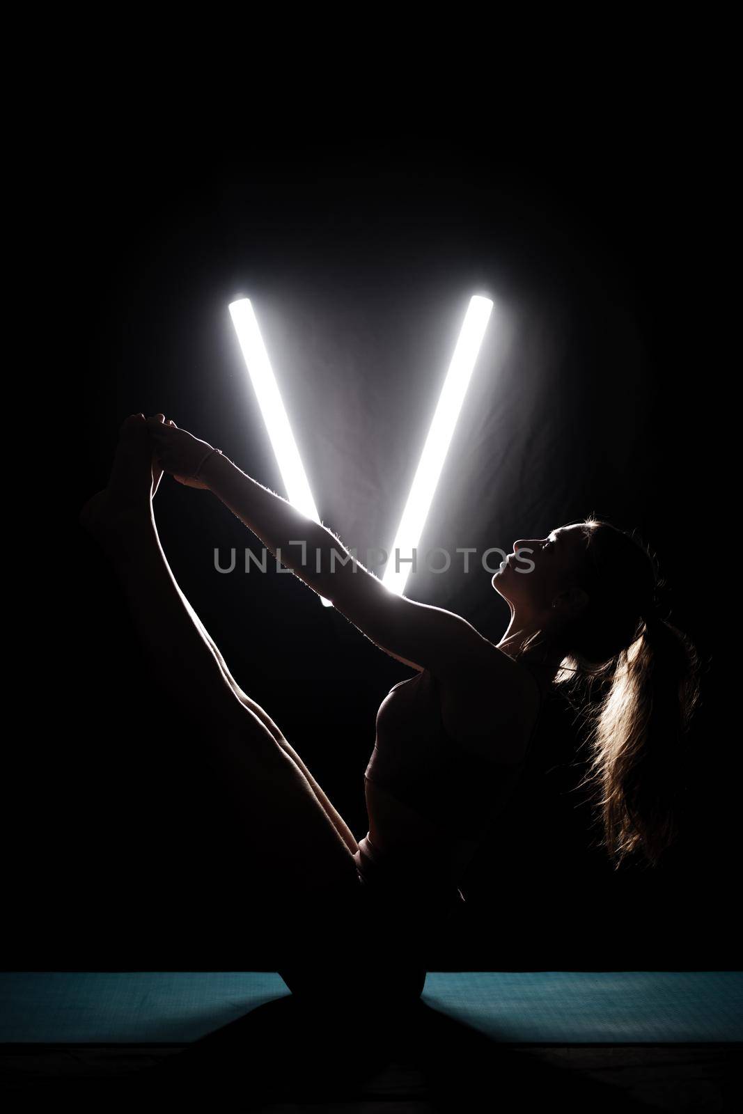 Fit woman practicing yoga poses. Silhouette girl doing exercise in studio against black background with v shaped white led tube light. No stress inner balance concept.. by kokimk