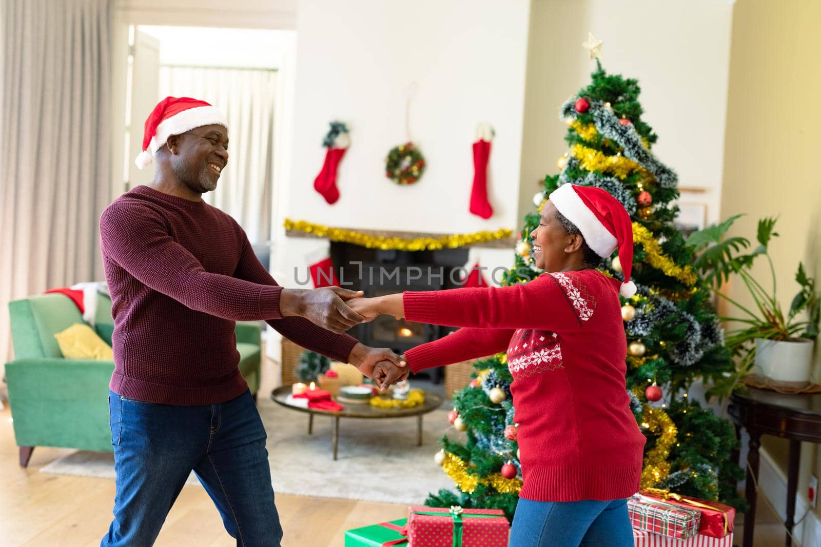 Happy african american senior couple in santa hats dancing at home at christmas time. retirement lifestyle and christmas festivities, celebrating at home.