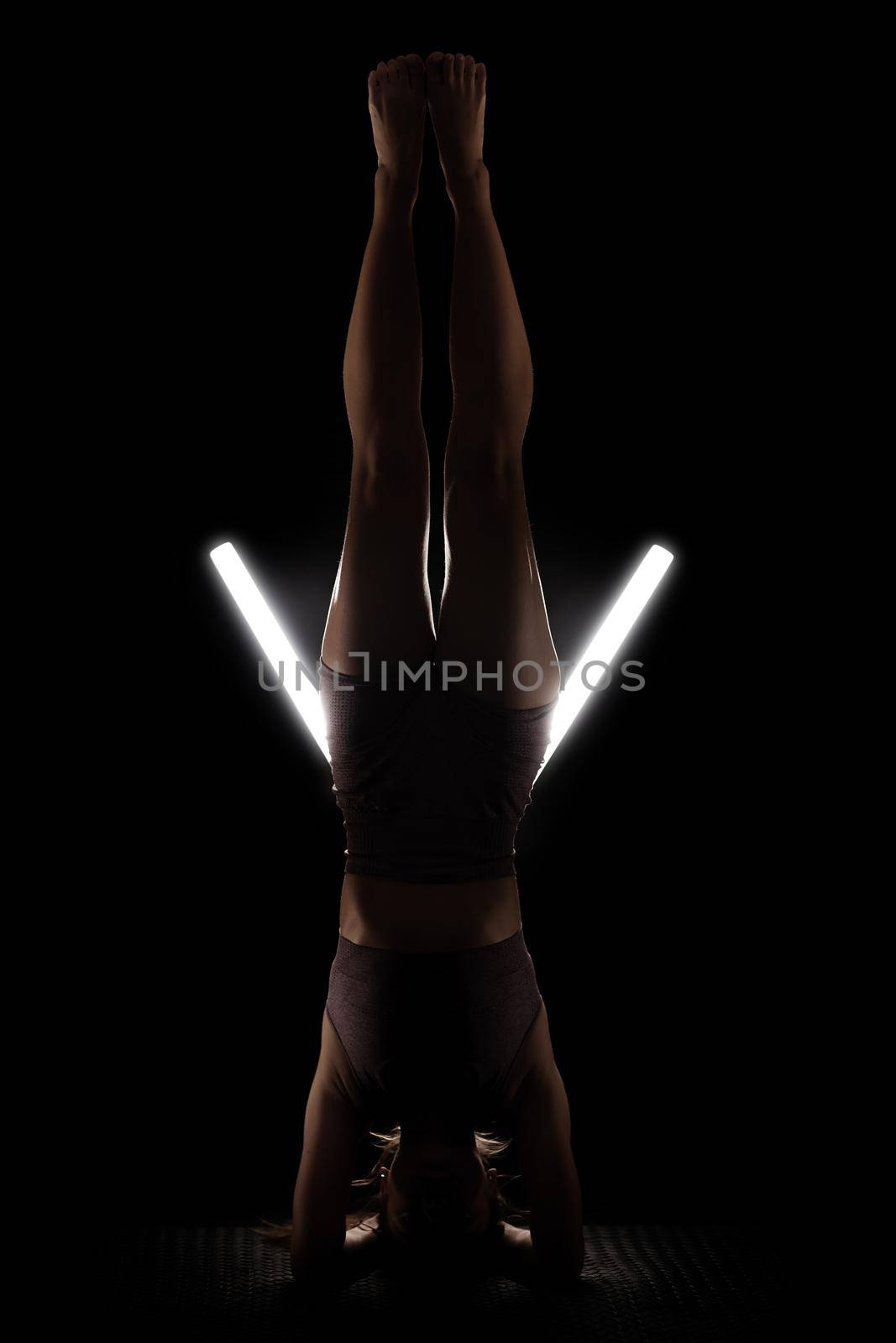 Fit woman practicing yoga poses. Silhouette girl doing exercise in studio against black background with v shaped white led tube light. No stress inner balance concept.. by kokimk