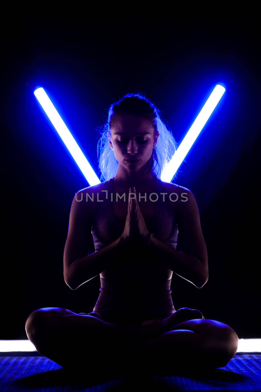 Fit woman practicing yoga poses. Silhouette girl doing exercise in studio against black background with v shaped neon blue or purple led tube light. by kokimk