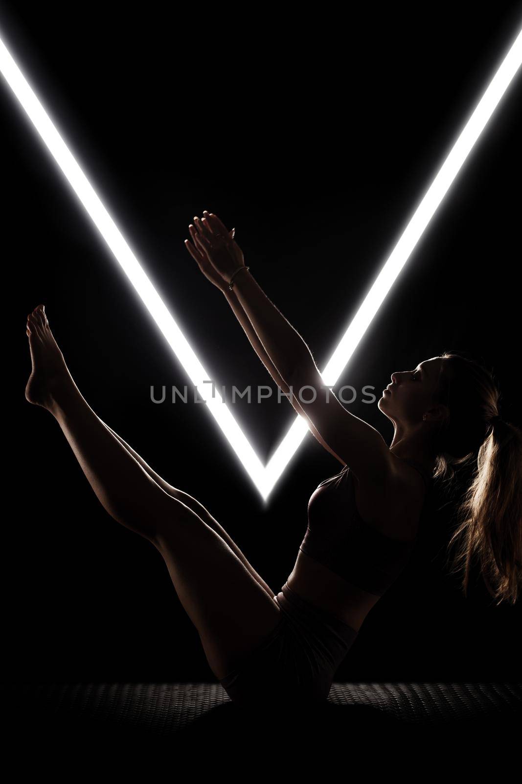 Fit woman practicing yoga poses. Silhouette girl doing exercise in studio against black background with v shaped white led tube light. No stress inner balance concept.