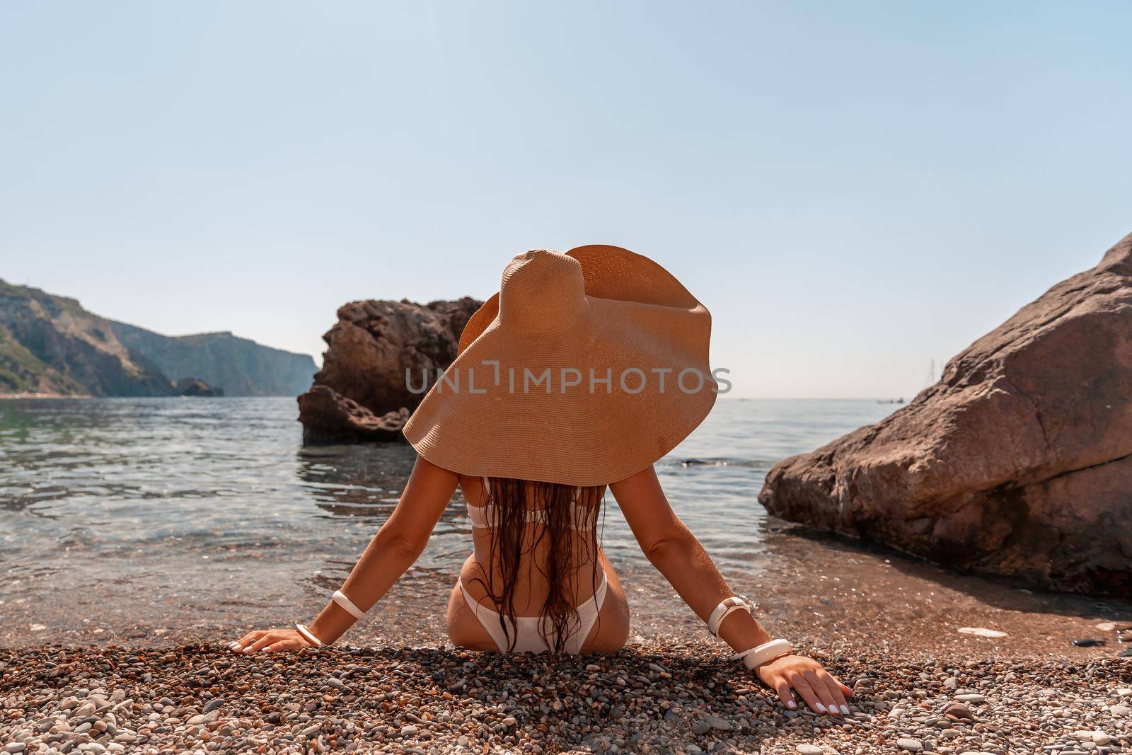 Tanned middle-aged woman with long hair and a white bathing suit. He sits on the seashore in a large sun hat with his back and looks at the sea. by Matiunina