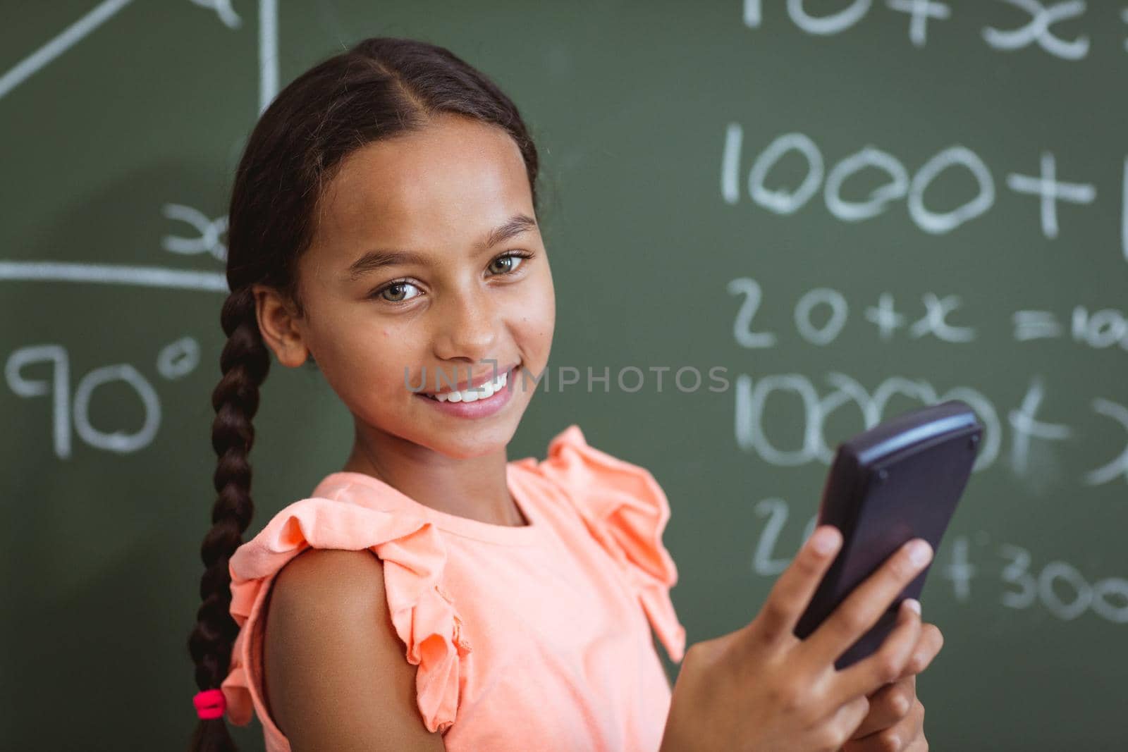 Portrait of smiling mixed race schoolgirl standing at chalkboard in classroom using smartphone. childhood, technology and education at elementary school.