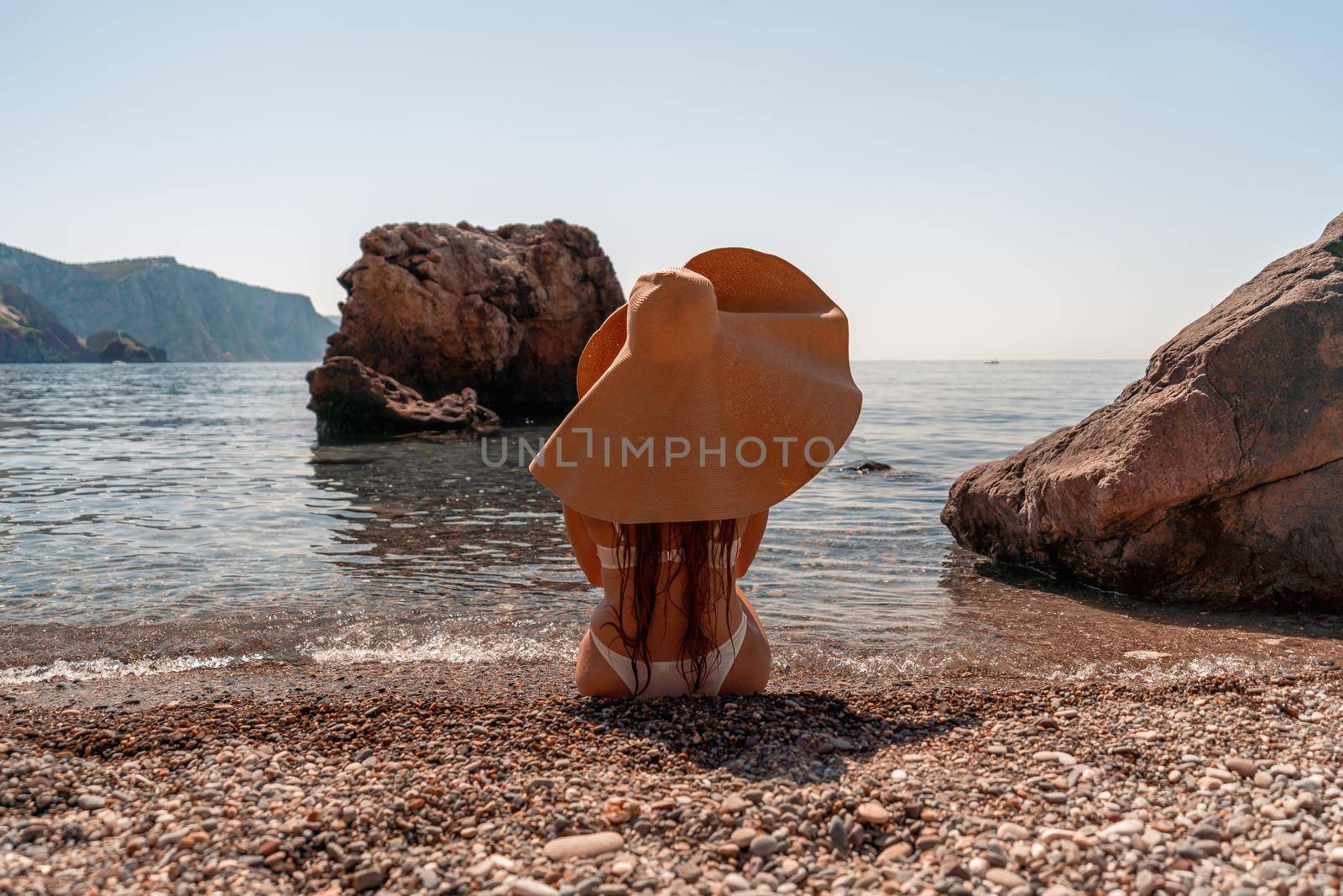 Tanned middle-aged woman with long hair and a white bathing suit. He sits on the seashore in a large sun hat with his back and looks at the sea. by Matiunina