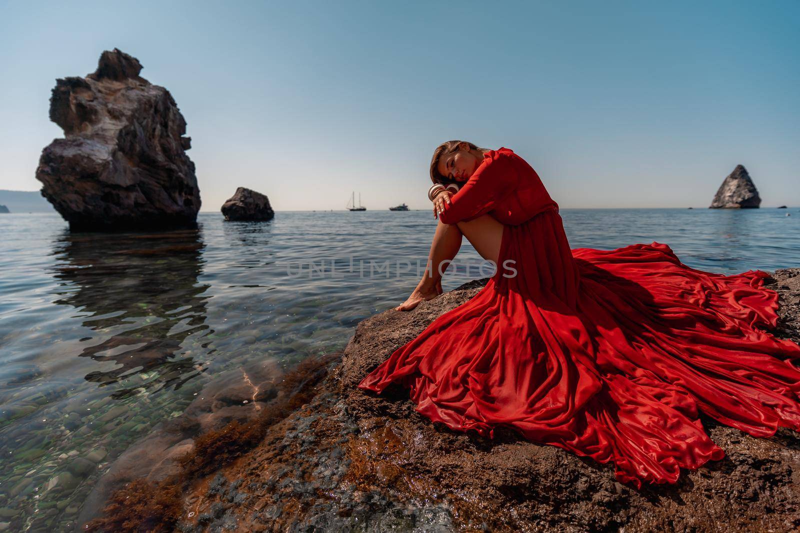 Beautiful sensual woman in a flying red dress and long hair, sitting on a rock above the beautiful sea in a large bay. by Matiunina