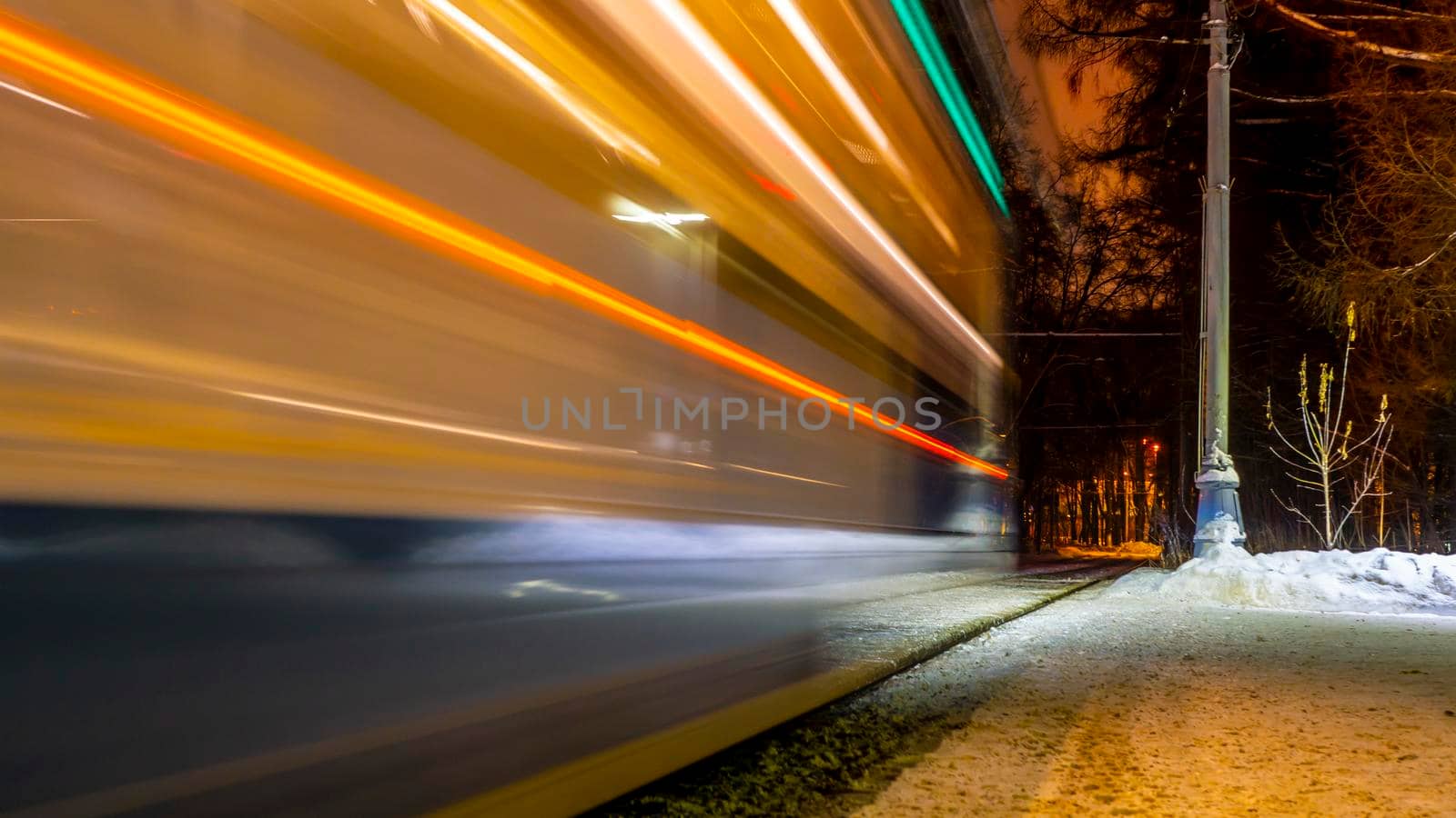 Public transportation in a city. Tram leaving the station, traffic lights, people, street, houses, overhead wires, rails. Movement and lights, long exposure.