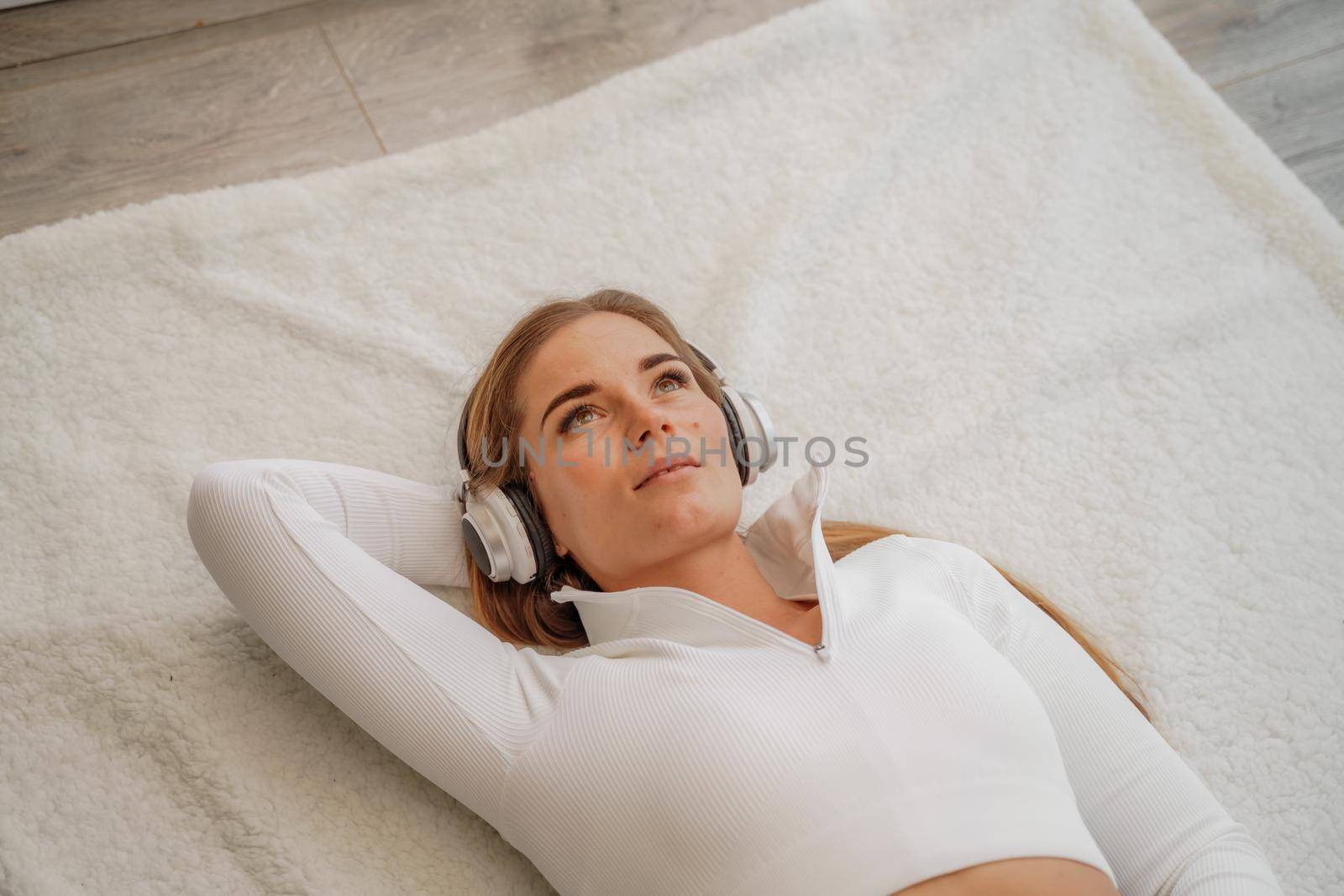 Top view portrait of relaxed woman listening to music with headphones lying on carpet at home. She is dressed in a White tracksuit. by Matiunina
