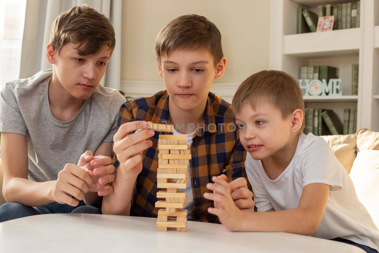 Three teen boy, brothers are playing a board game made of wooden rectangular blocks, pulling pieces out of the tower. by Zakharova