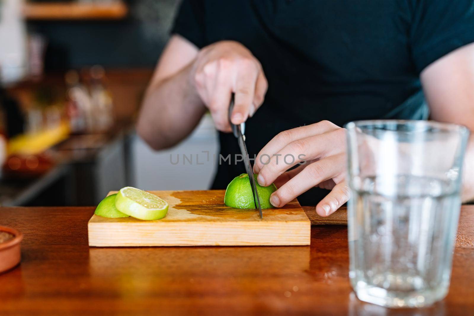 Young and modern waiter, with long dark hair, dressed in black polo shirt, cutting slices of citrus fruit on a wooden board, to prepare a cocktail. Waiter preparing a cocktail. Cocktail glass with ice cubes. Gin and tonic. Bar full of cocktail ingredients. Dark background and dramatic lighting.