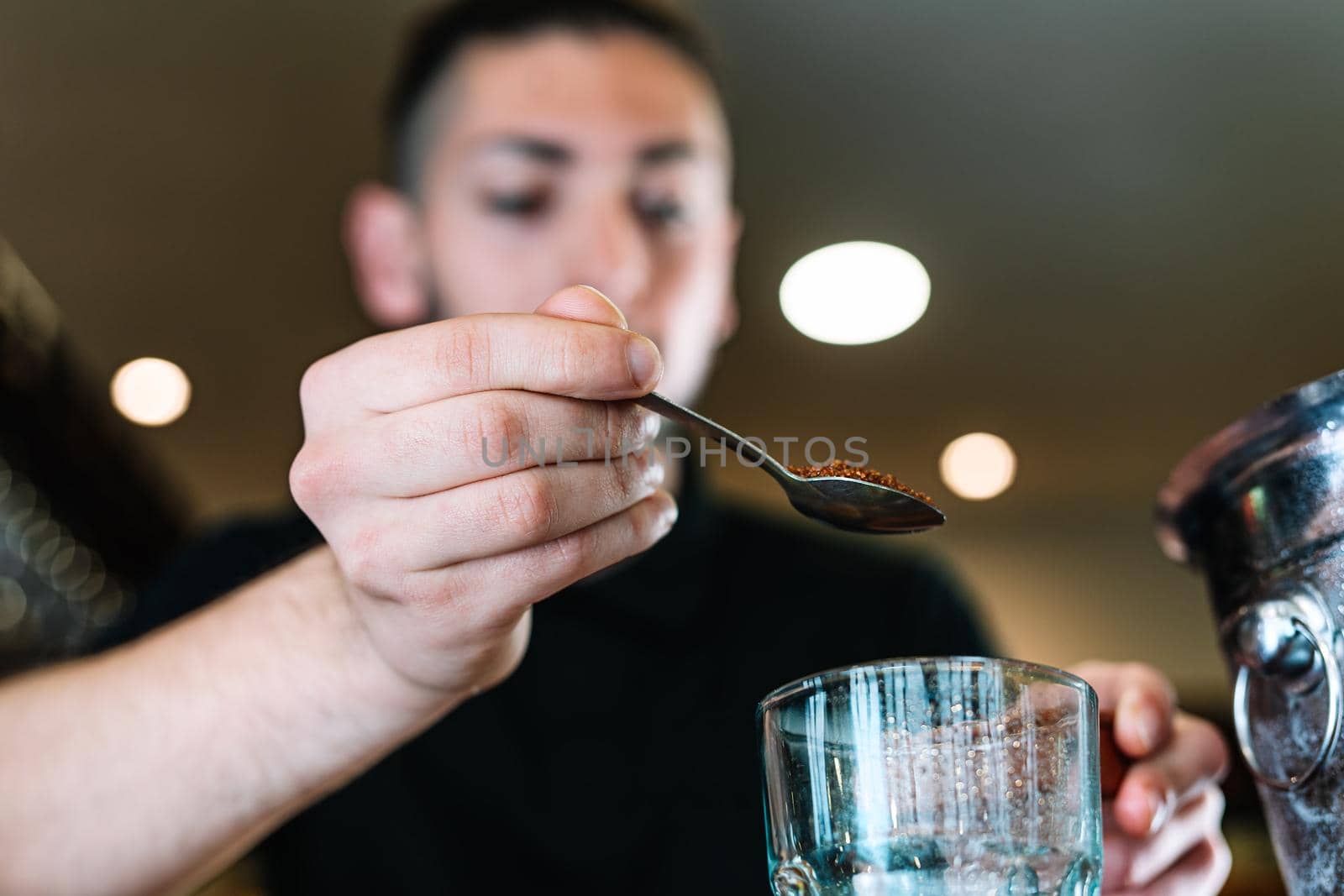 Young and modern waiter, with long dark hair, dressed in black polo shirt, adding brown sugar in a large crystal glass to prepare a cocktail. Waiter preparing a cocktail. Cocktail glass with ice cubes. Mojito. Bar full of cocktail ingredients. Dark background and dramatic lighting. low angle view