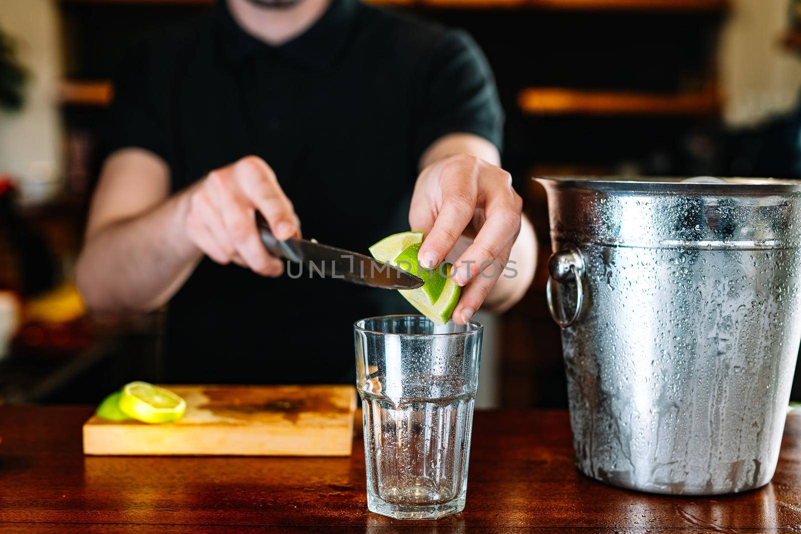 Young and modern waiter, with long dark hair, dressed in black polo shirt, squeezing slices of citrus fruit to prepare a cocktail. Waiter preparing a cocktail. Cocktail glass with ice cubes. Gin and tonic. Bar full of cocktail ingredients. Dark background and dramatic lighting.