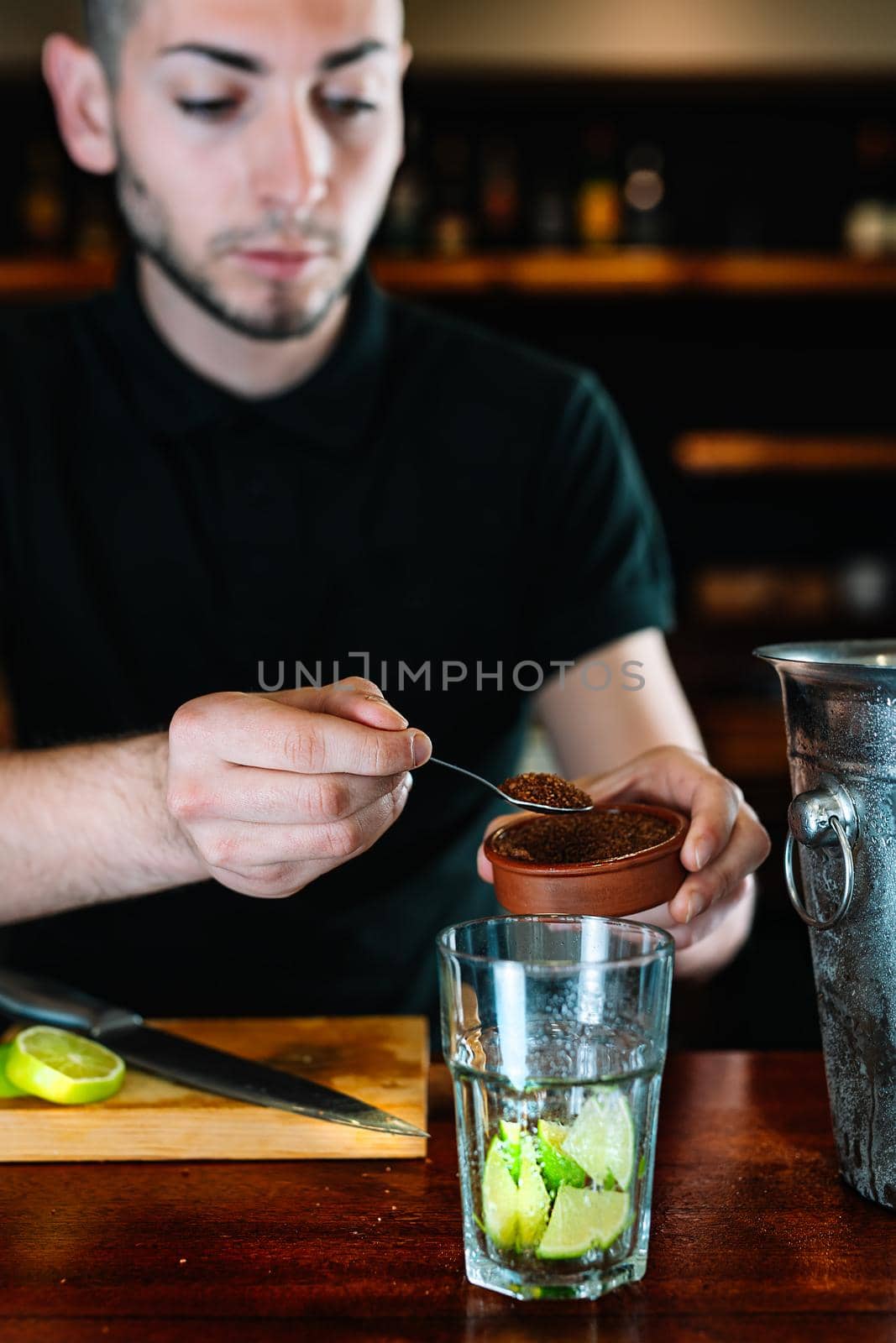 Young and modern waiter, with long dark hair, dressed in black polo shirt, adding brown sugar in a large crystal glass to prepare a cocktail. Waiter preparing a cocktail. Cocktail glass with ice cubes. Mojito. Bar full of cocktail ingredients. Dark background and dramatic lighting. Vertical