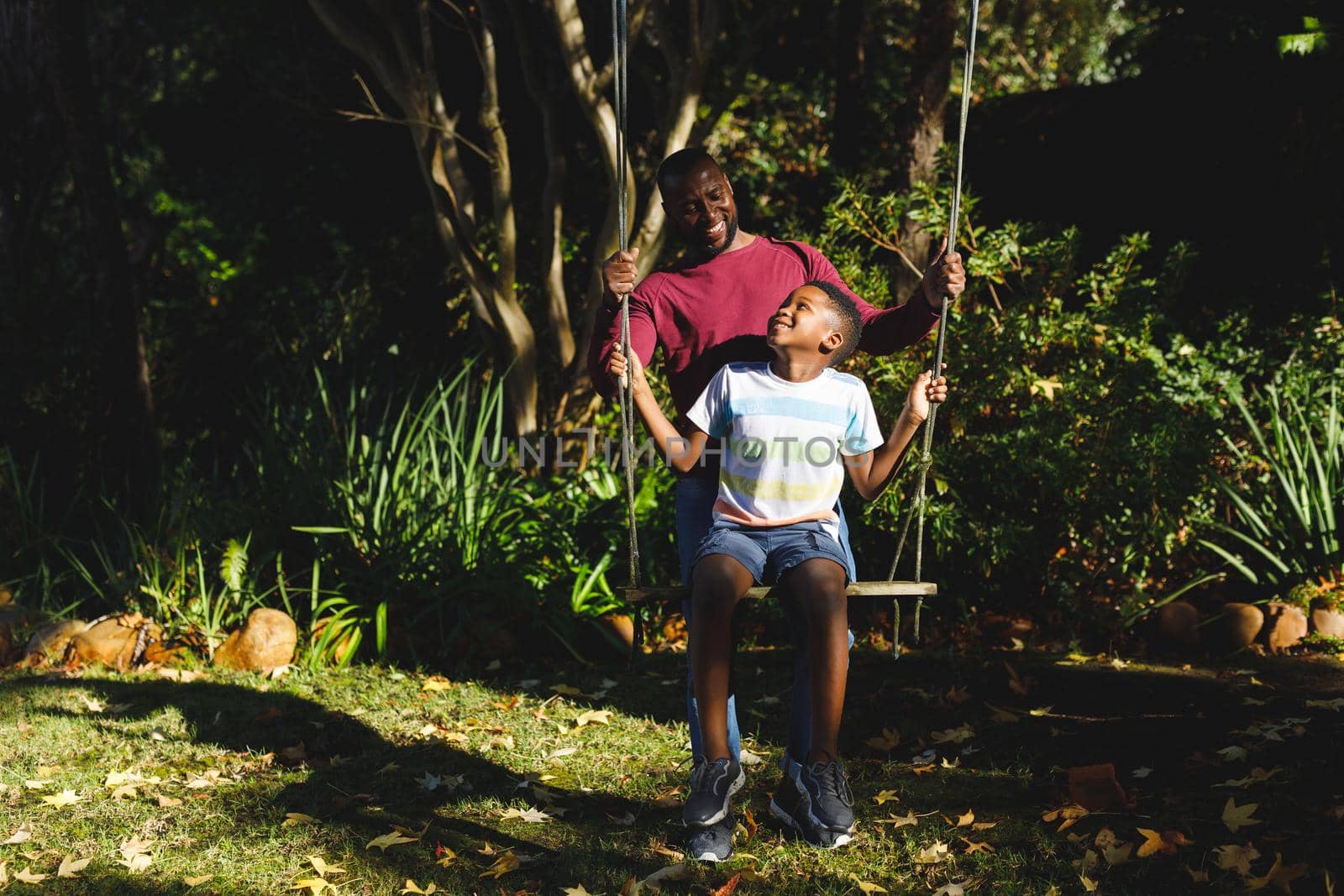 African american father with son having fun and playing on swing in garden. family spending time at home.