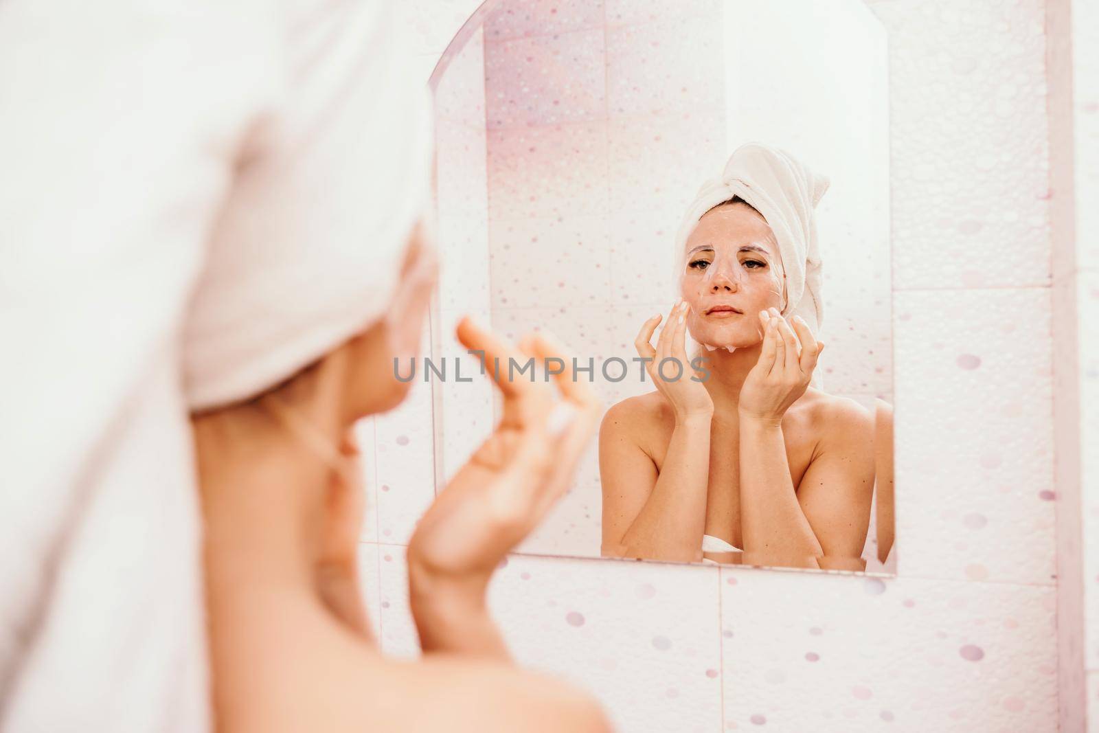 Young beautiful woman using a moisturizing facial mask after taking a bath. Pretty attractive girl in a towel on her head stands in front of a mirror in a home bathroom. Daily hygiene and skin care.