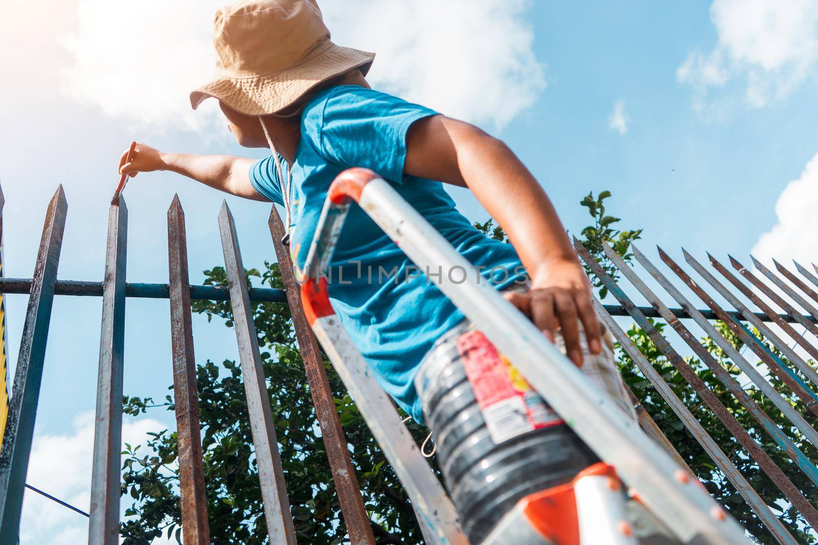 Latin woman dressed in shorts, t-shirt and hat, standing on a ladder standing to paint the highest part of a metal fence outdoors.