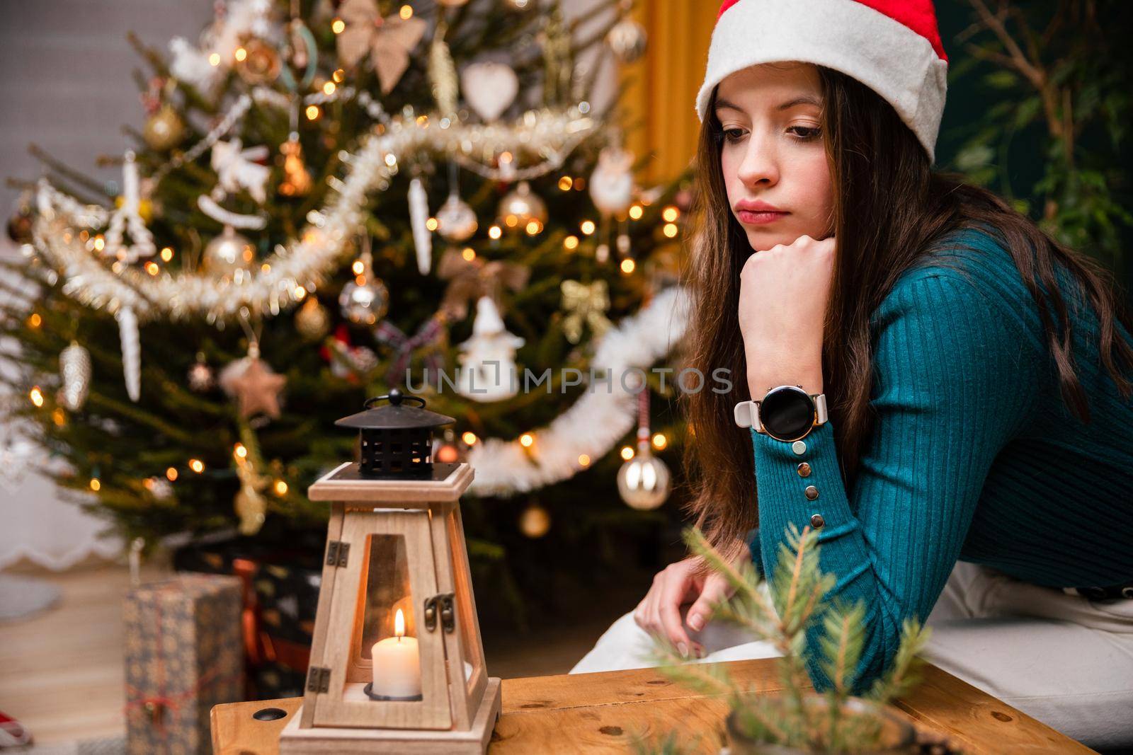 A teenage girl is spending Christmas alone. A sad girl sits at a table wearing a Santa hat against the background of a Christmas tree. Background with bokeh effect.