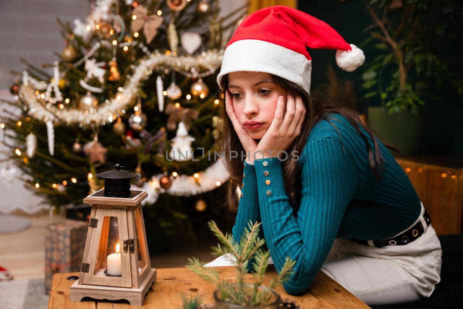 A teenage girl is spending Christmas alone. A sad girl sits at a table wearing a Santa hat against the background of a Christmas tree. Background with bokeh effect.