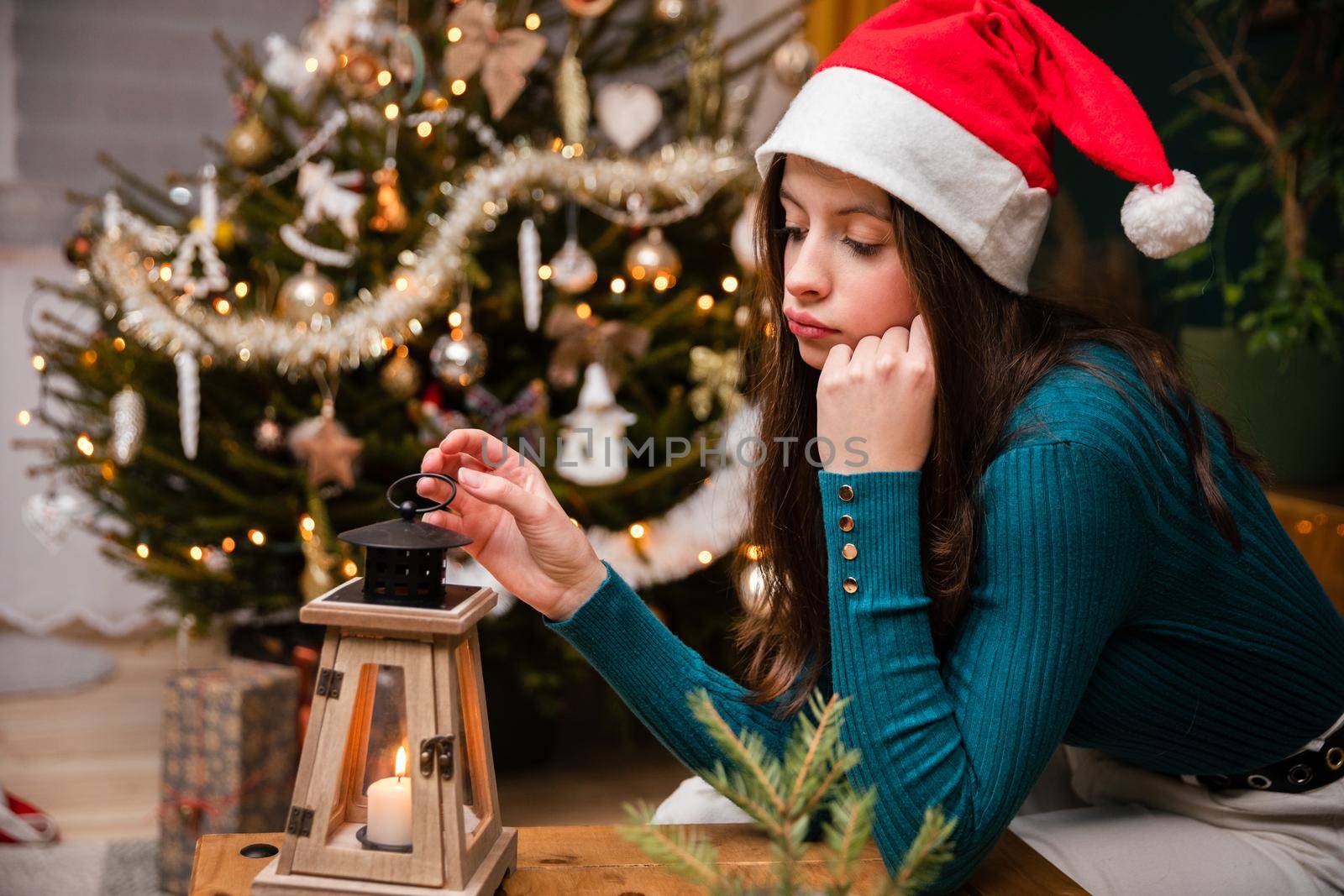 A teenage girl is spending Christmas alone. A sad girl sits at a table wearing a Santa hat against the background of a Christmas tree. Background with bokeh effect.