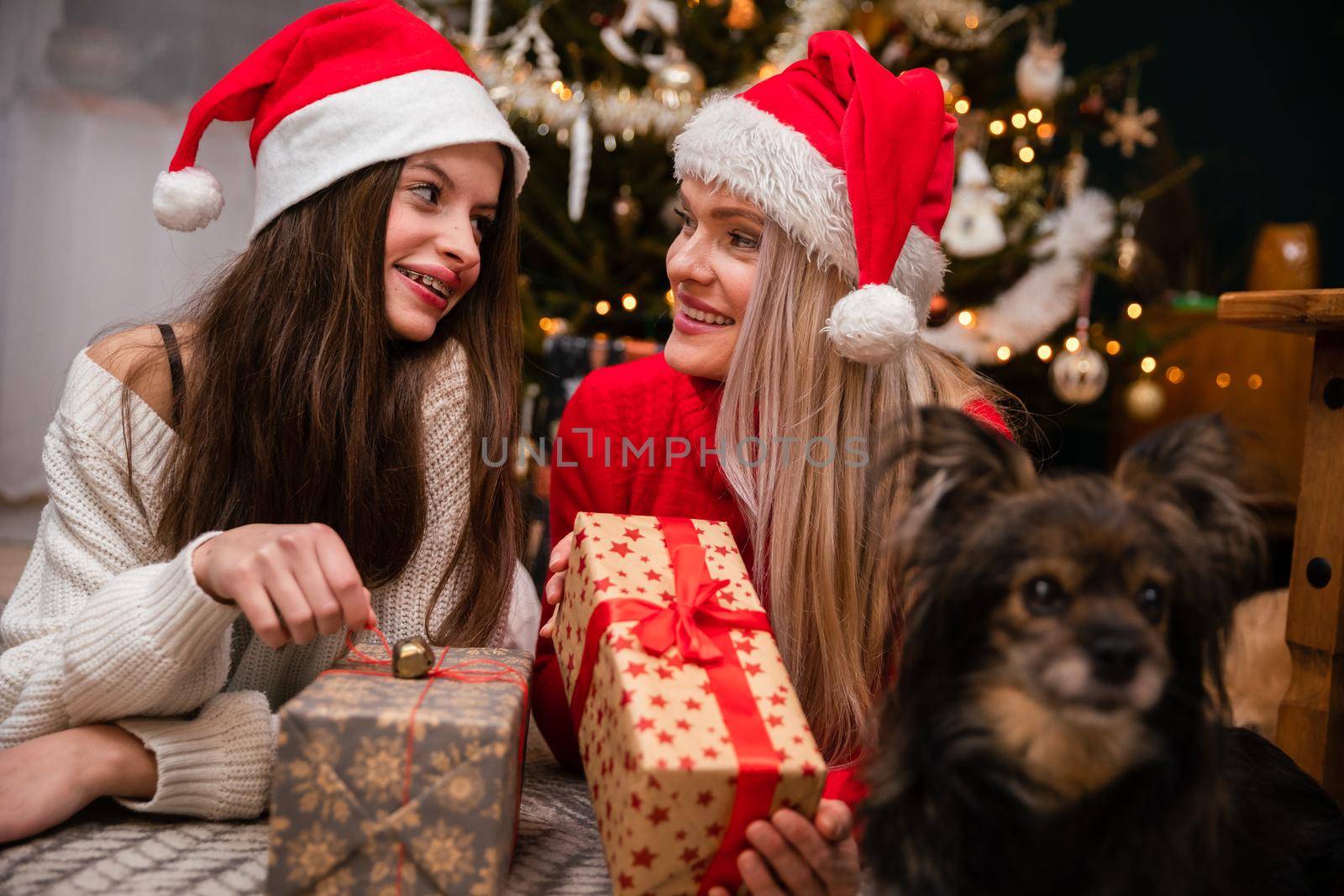 Mother and daughter are lying smiling on the floor with gifts in their hands and looking into each other's eyes. A mongrel dog is lying next to the girls.