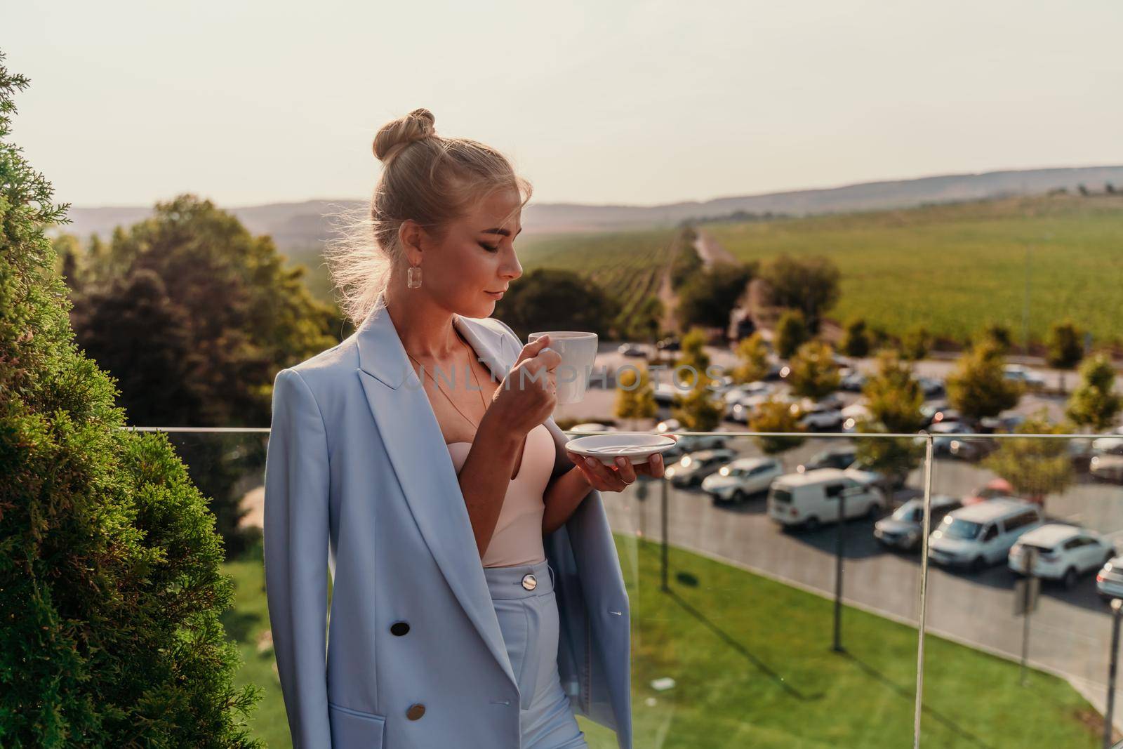 A middle-aged woman stands in a street cafe overlooking the mountains at sunset. She is wearing a blue jacket and drinking coffee while admiring nature. Travel and vacation concept