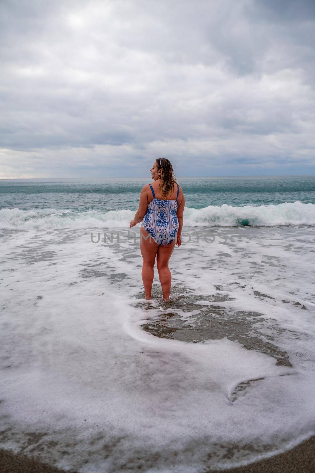 A plump woman in a bathing suit enters the water during the surf. Alone on the beach, Gray sky in the clouds, swimming in winter