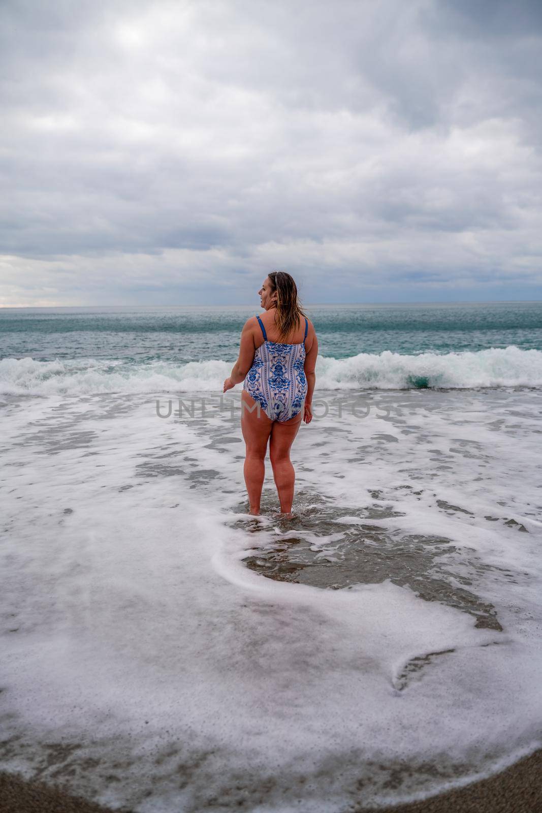 A plump woman in a bathing suit enters the water during the surf. Alone on the beach, Gray sky in the clouds, swimming in winter. by Matiunina