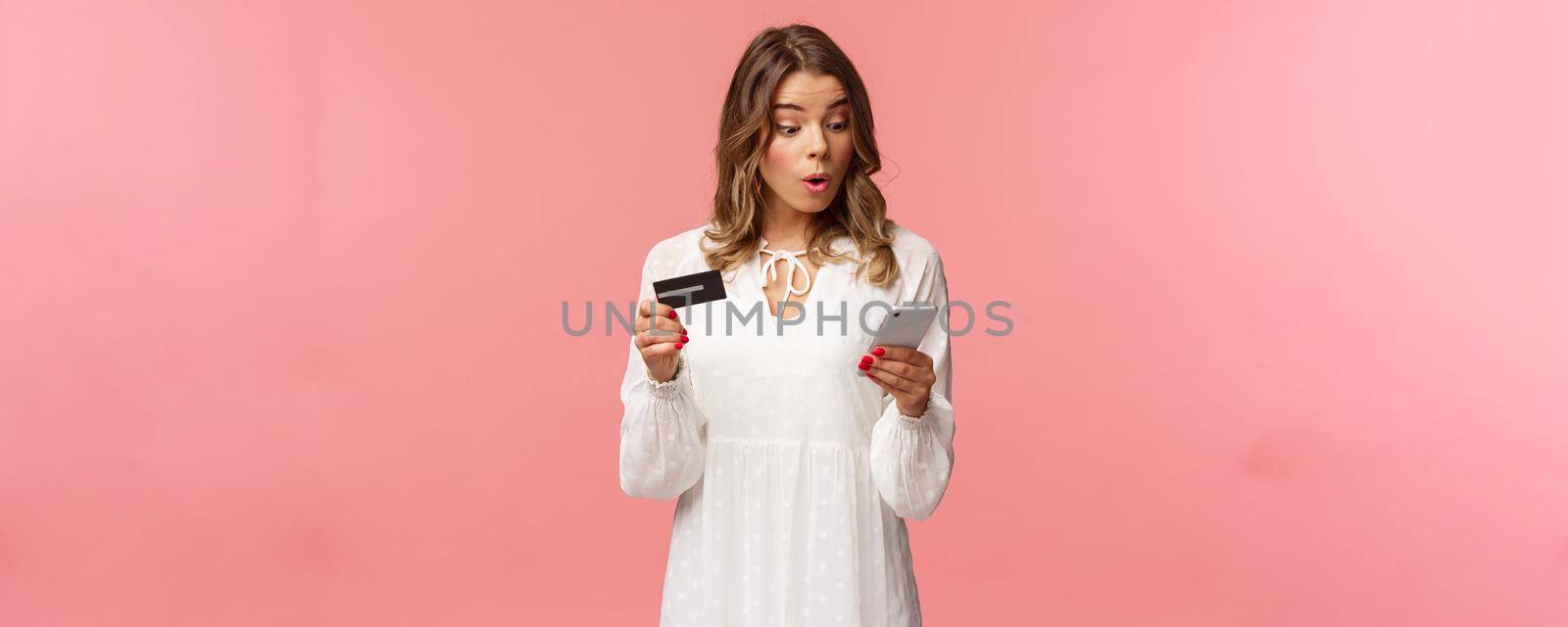 Excited and intrigued young blond girl in white dress, hurry up and make order on online shopping site before discount ends, enter credit card digit numbers to mobile phone, pink background.