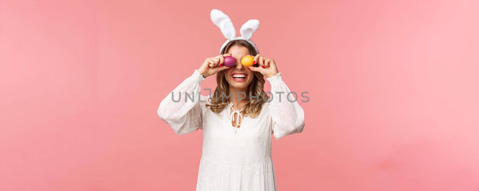 Holidays, spring and party concept. Portrait of lovely, tender smiling woman in rabbit ears and white dress celebrating Easter day, holding painted eggs on eyes and grinning, pink background.