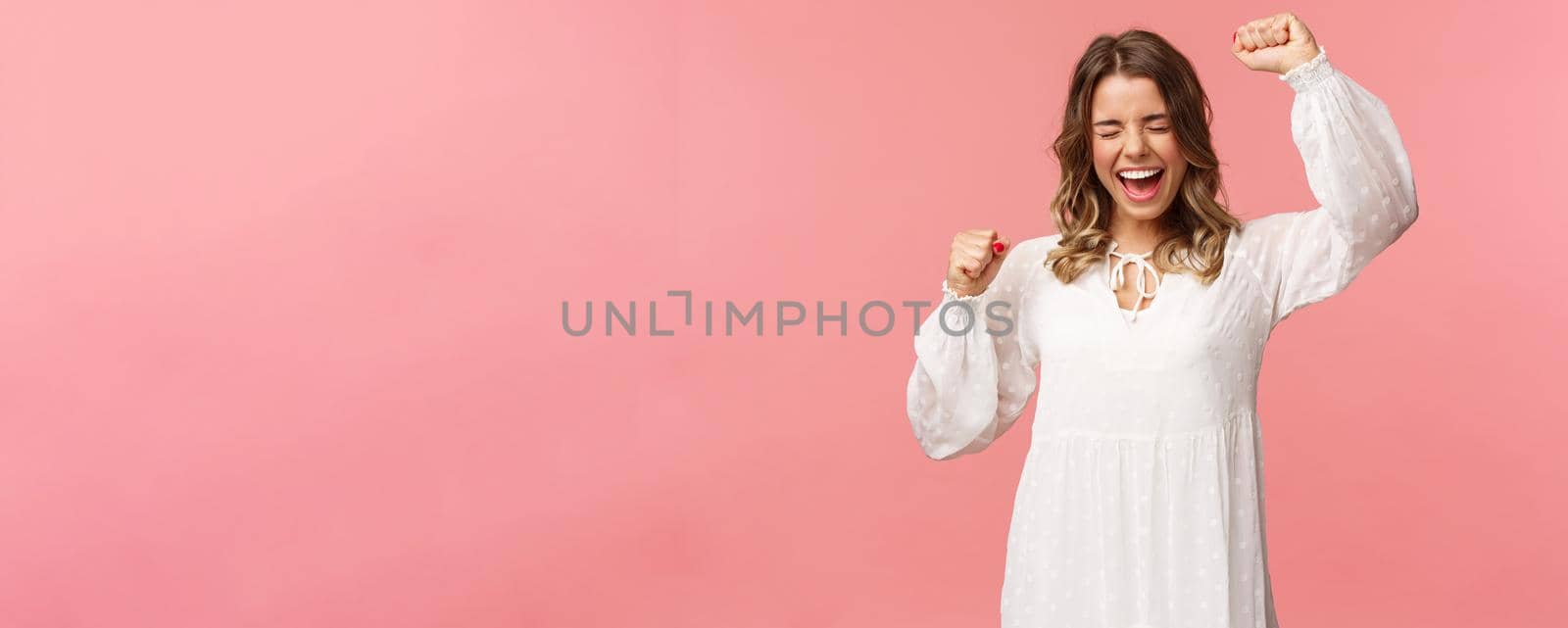 Portrait of beautiful tender young blond girl in white dress, raising hands up in hooray, yes or victory, shouting relieved, scream from happiness, triumphing, winning prize, become champion.