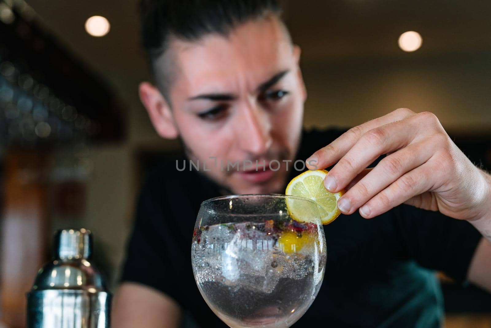 Young and modern waiter, with long dark hair, dressed in black polo shirt, decorating with a slice of citrus fruit a cocktail in a crystal glass with ice for a cocktail. Waiter preparing a cocktail. Cocktail glass with ice cubes. Gin and tonic. Bar full of cocktail ingredients. Dark background and dramatic lighting.