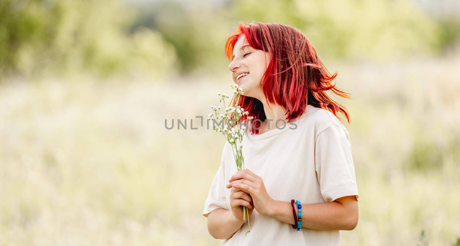 Teen girl with red hair at the nature smiling with bouquet in hands. Pretty young female in the sunny field